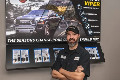 Bearded man in black uniform stands arms crossed before a Viper security product display