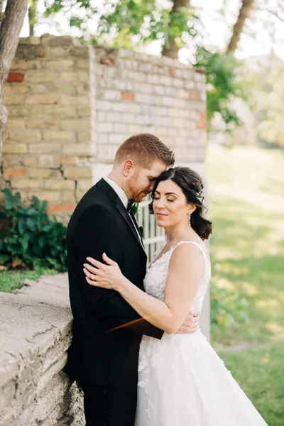 Bride in white lace gown embracing groom in black tuxedo outdoors near a stone wall