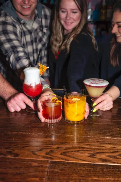 Three people holding four colorful cocktails on a wooden bar top