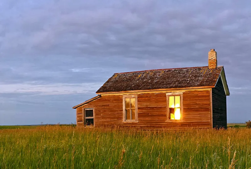Weathered wooden farmhouse on open prairie at dusk, warm light glowing through windows