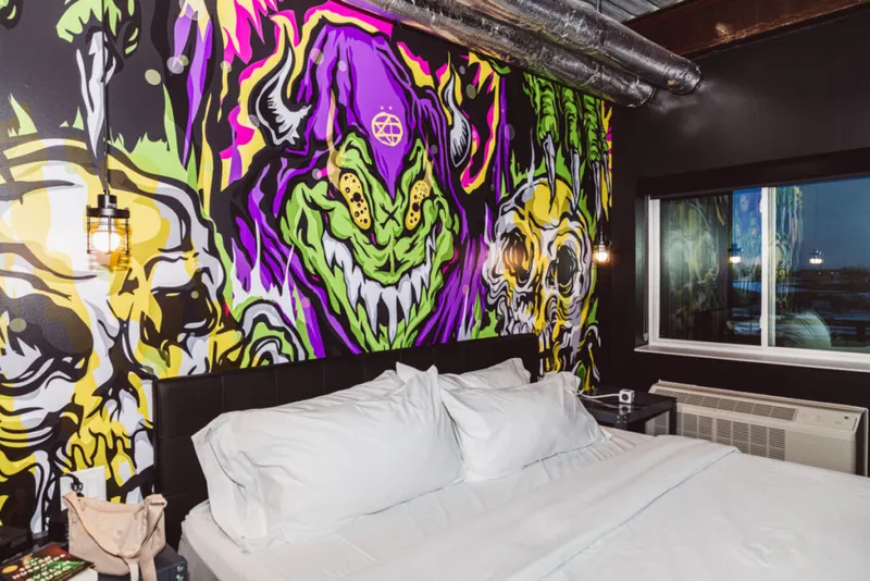 Hotel room with white bedding backed by a bold graffiti mural featuring snarling monster faces in purple, green, and yellow