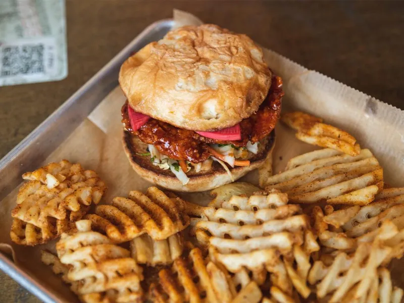 Saucy fried chicken sandwich with slaw on a biscuit, served on a metal tray with seasoned waffle fries