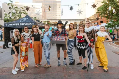 Group of nine colorfully costumed performers posing outdoors near a Drekker Brewing tent