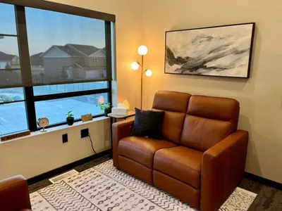 Cozy therapy room with a brown leather loveseat, floor lamp, and abstract wall art near a large window