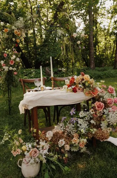 Outdoor sweetheart table draped in white linen, surrounded by lush arrangements of roses and wildflowers with tall candles