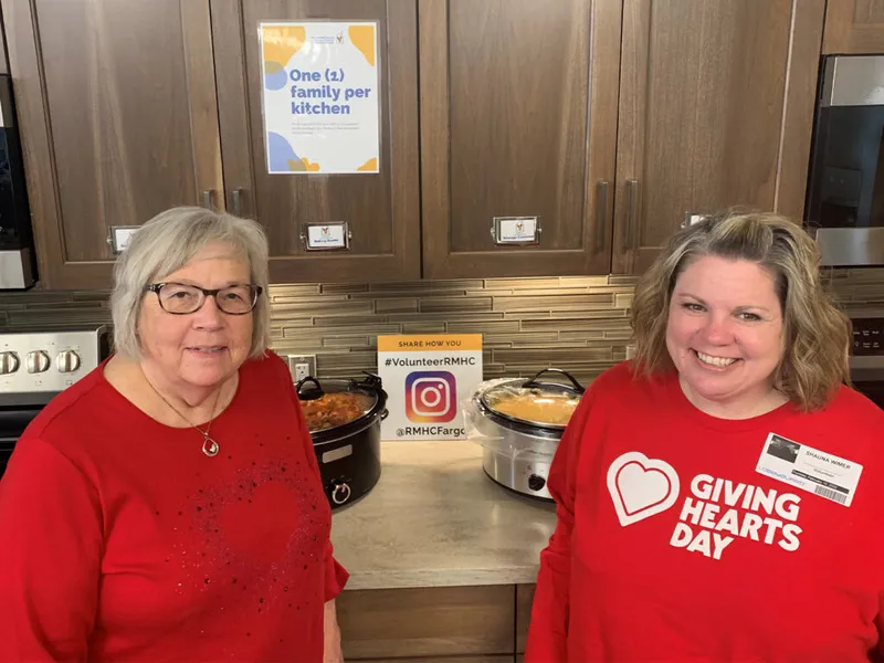 Two women in red sweatshirts smile beside slow cookers in a Ronald McDonald House kitchen