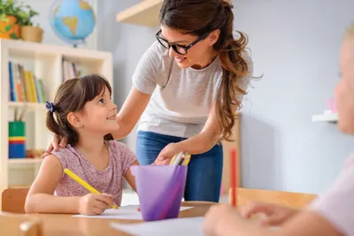 Teacher leaning down to smile at a young girl drawing at a classroom desk