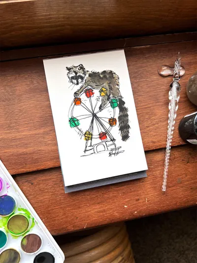 Small ink-and-watercolor illustration of a raccoon atop a Ferris wheel, beside a glass dip pen and watercolor palette
