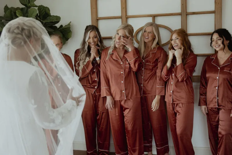 Veiled bride facing bridesmaids in matching rust satin pajamas reacting with emotional smiles