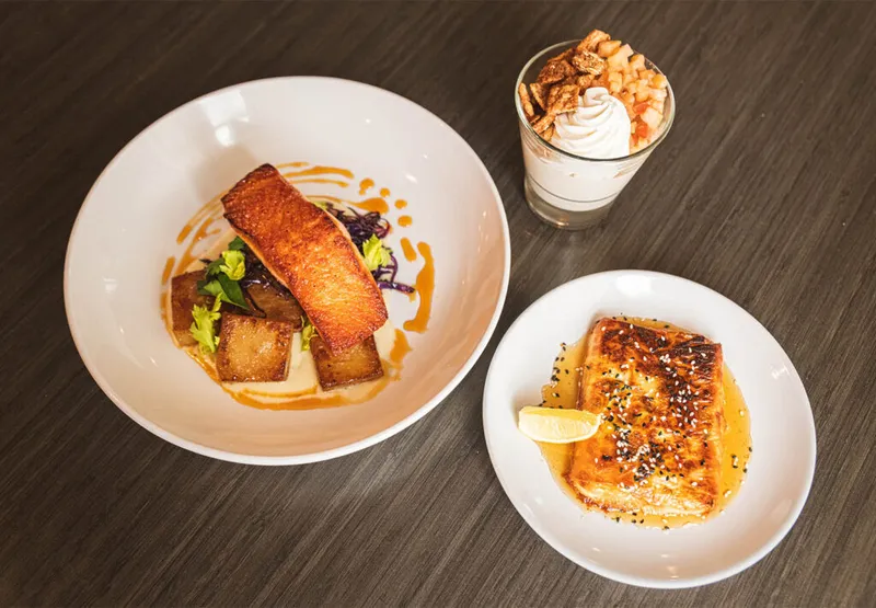 Overhead view of seared salmon bowl, sesame-crusted halloumi plate, and a whipped cream dessert glass on a dark wood table