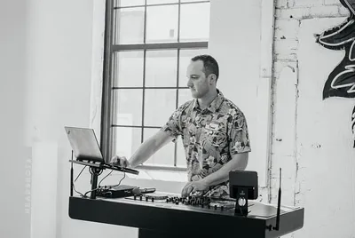 Man in floral shirt DJing at a turntable setup near a large window