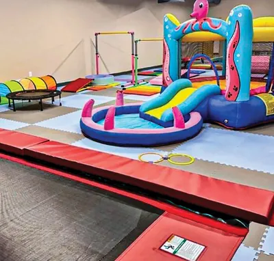 Colorful indoor play area with inflatable bounce house, slide, foam mats, and gymnastics bars