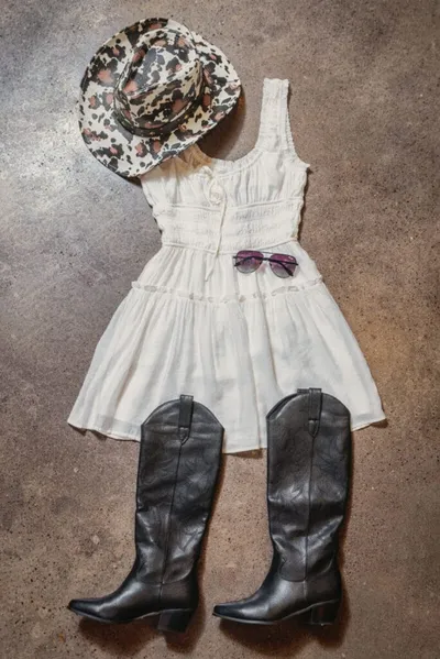 Flat lay of white sundress, black cowboy boots, floral hat, and aviator sunglasses