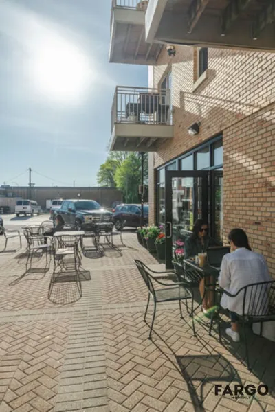 Two women sitting at a sunny brick patio outside a tan brick building with black-framed windows