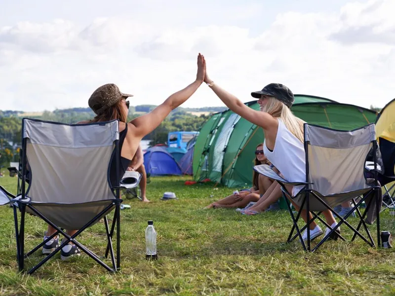 Two women in caps high-five from camping chairs at an outdoor music festival