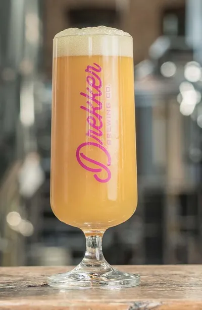 Foamy golden hazy beer in a stemmed Drekker Brewing Co. glass on a wood surface