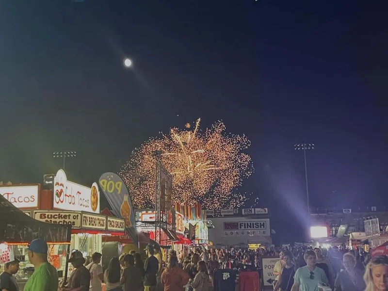 Fireworks burst over a crowded fairground midway at night with food vendor signs lit up below
