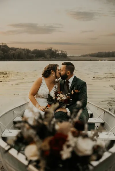 Bride and groom kissing in a rowboat on a calm lake at dusk, bouquet in foreground