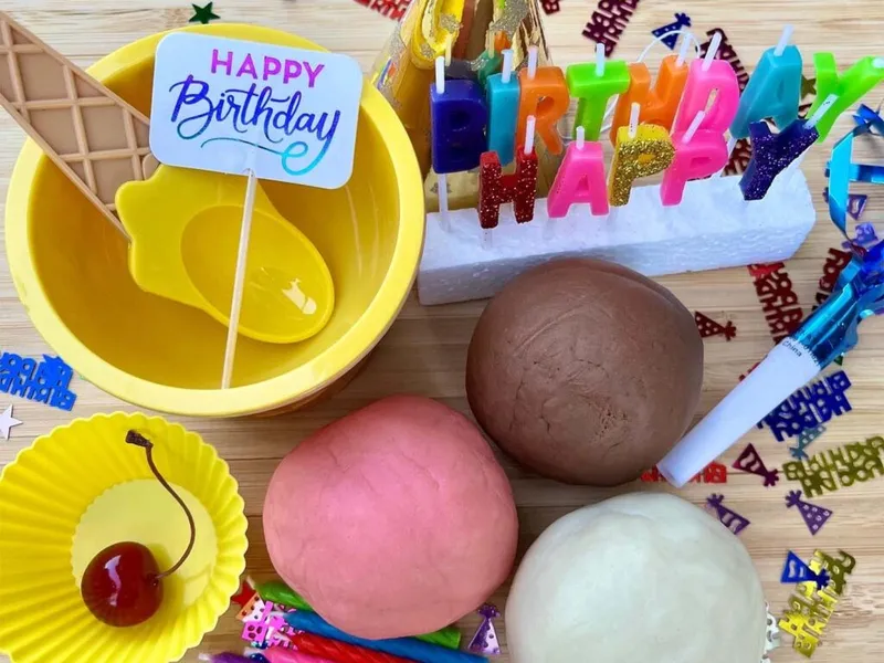 Birthday-themed play dough kit with colorful candles, yellow bowl, cherry, and three dough balls on wood surface