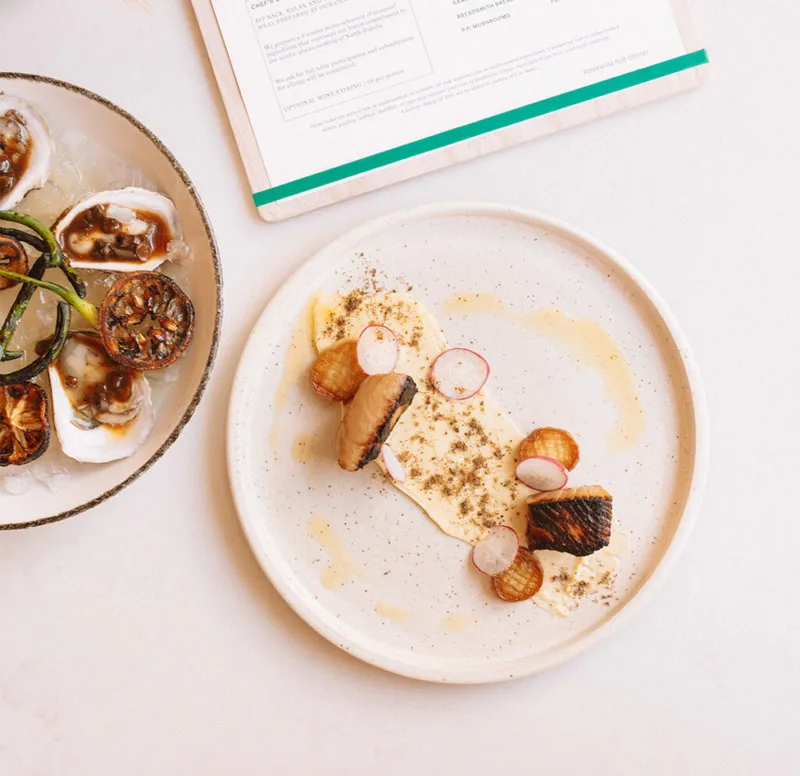 Plated seared fish with sliced radish and cream sauce beside oysters on ice, with a menu