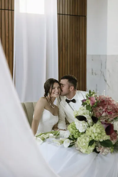 Groom in white suit kissing bride's cheek at a floral-adorned sweetheart table