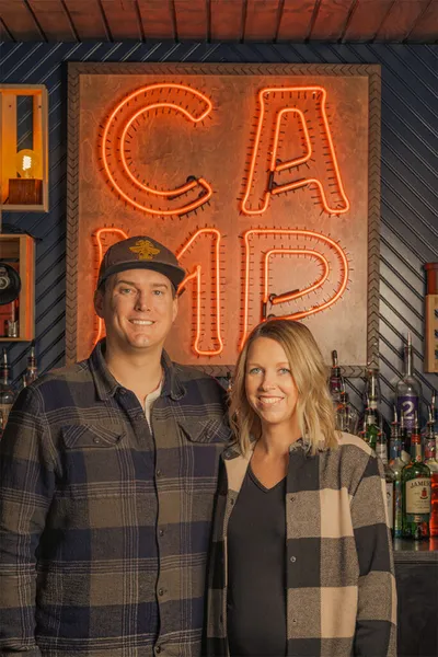 Man and woman smiling in front of an orange neon 'CAMP' sign above a bar