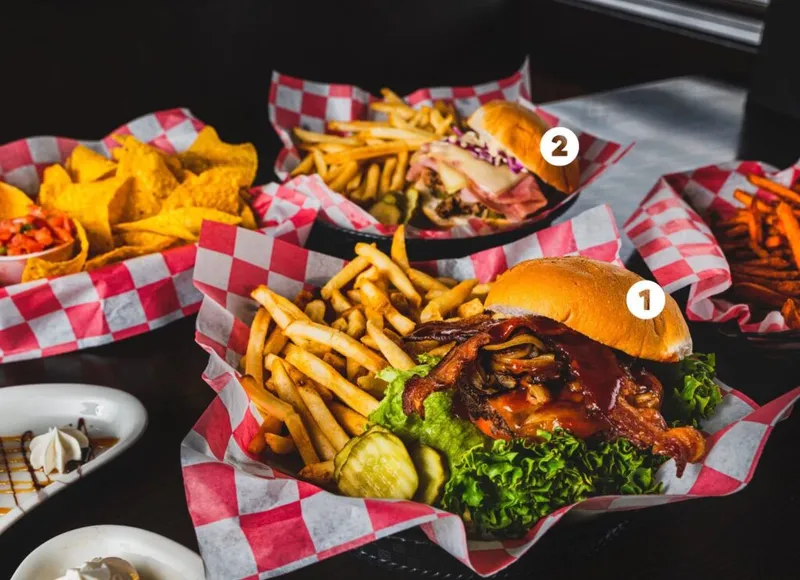 Burger with bacon and fries in a red checkered basket alongside nachos and a second sandwich