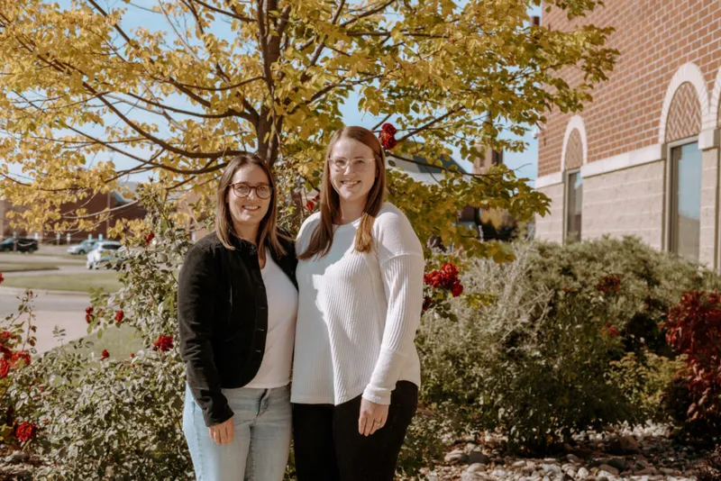 Two smiling women in glasses standing outdoors near red roses and an autumn tree