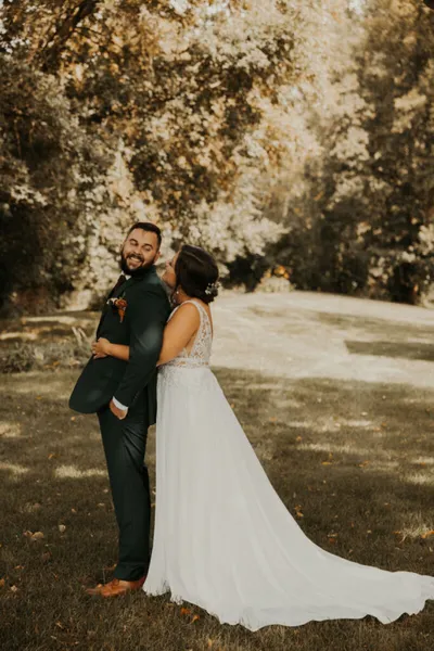 Bride in white gown embraces groom in dark suit outdoors among autumn trees