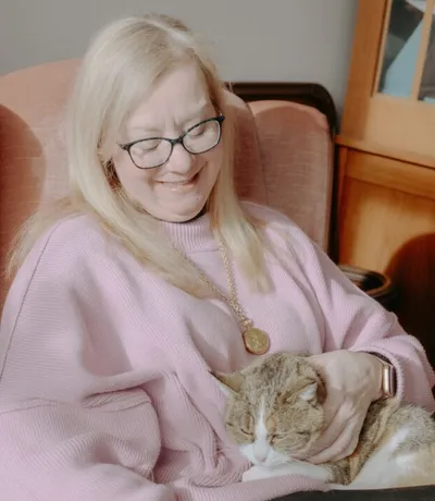 Smiling woman with long blonde hair and glasses petting a tabby cat in a pink armchair