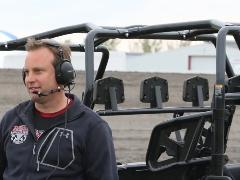Man wearing a racing jacket and headset mic beside a roll-cage UTV on a dirt track
