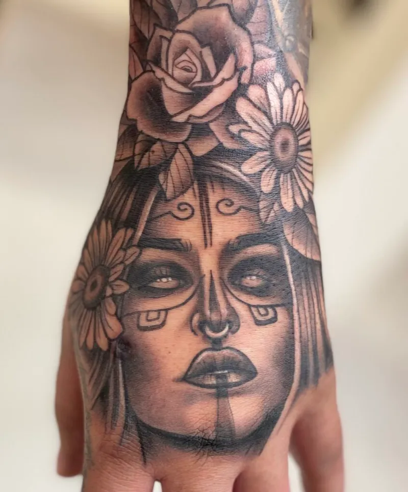 Black and grey hand tattoo of a woman's face with floral headdress and ornate facial markings