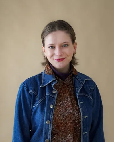 Young woman in denim jacket and patterned turtleneck smiling against beige background