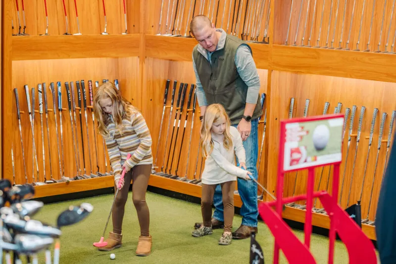 Man supervising two young girls putting on an indoor green surrounded by golf club displays