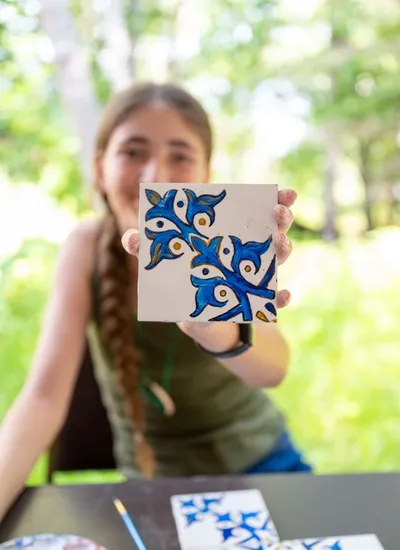 Girl with braided hair holds up a hand-painted blue floral tile outdoors