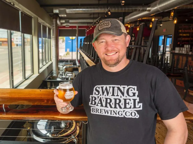Smiling man in a Swing Barrel Brewing Co. shirt and cap holding a glass of golden beer inside a taproom