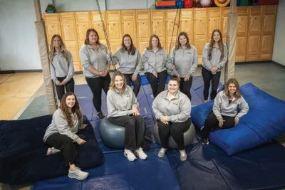 Group of ten women in matching gray sweatshirts posing in a therapy gym with blue mats