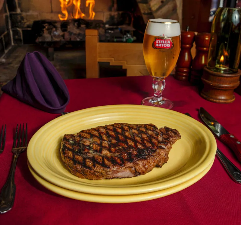 Grilled steak on a yellow plate beside a Stella Artois beer, red tablecloth, fireplace behind