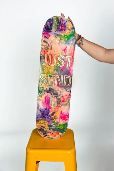 Hand holding a colorfully painted skateboard deck reading 'Just Send It' on a yellow stool