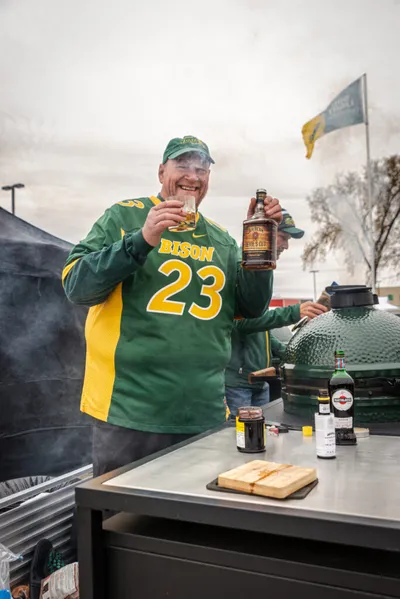 Smiling man in green Bison #23 jersey holding a whiskey bottle and glass at an outdoor grill setup
