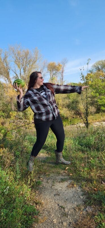 Woman in plaid shirt and rubber boots poised to throw a small watermelon outdoors