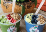 Two colorful smoothie bowls topped with granola and fruit beside a coconut drink