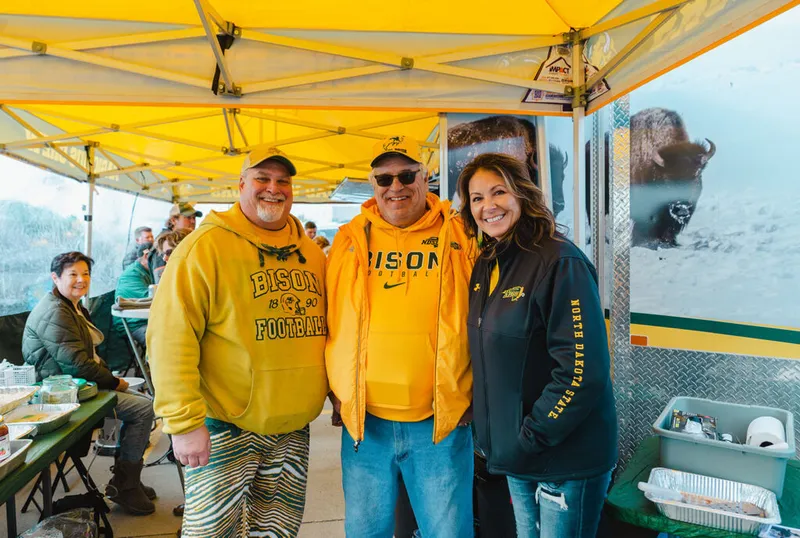Three smiling fans in NDSU Bison gear pose under a yellow canopy at a tailgate