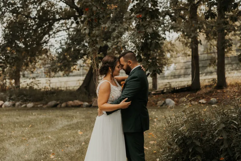 Bride in white gown and groom in dark suit embrace forehead-to-forehead beneath a large tree outdoors
