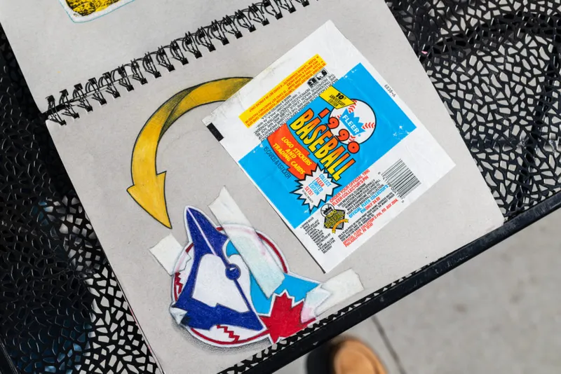 Sketchbook open on a metal table with a Blue Jays logo drawing, arrow sketch, and a vintage Fleer Baseball card wrapper