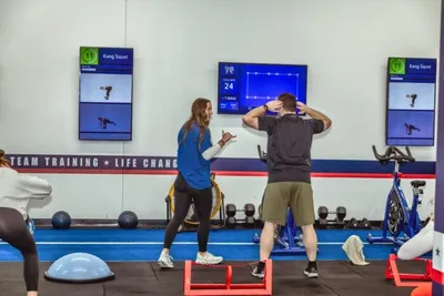 Trainer with clipboard coaching a man doing squats in a group fitness studio