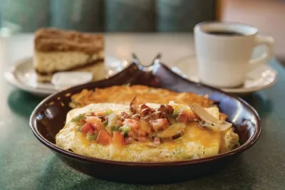 Cheese omelette topped with tomatoes and bacon in a brown bowl, with cake and coffee behind