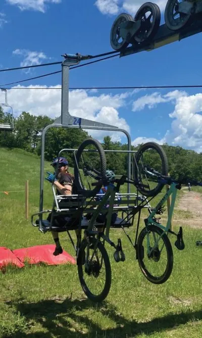 Helmeted rider on a ski chairlift carrying two mountain bikes up a grassy summer hill