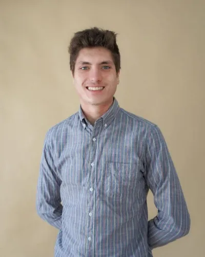 Young man with dark hair smiling, wearing a striped button-down shirt, against a beige background