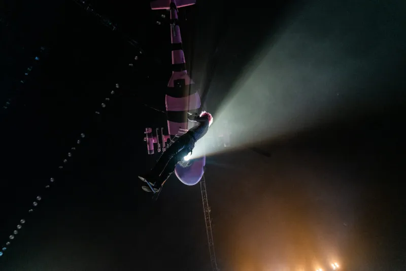 A performer descends from the rafters in a spotlight beam above a darkened arena stage